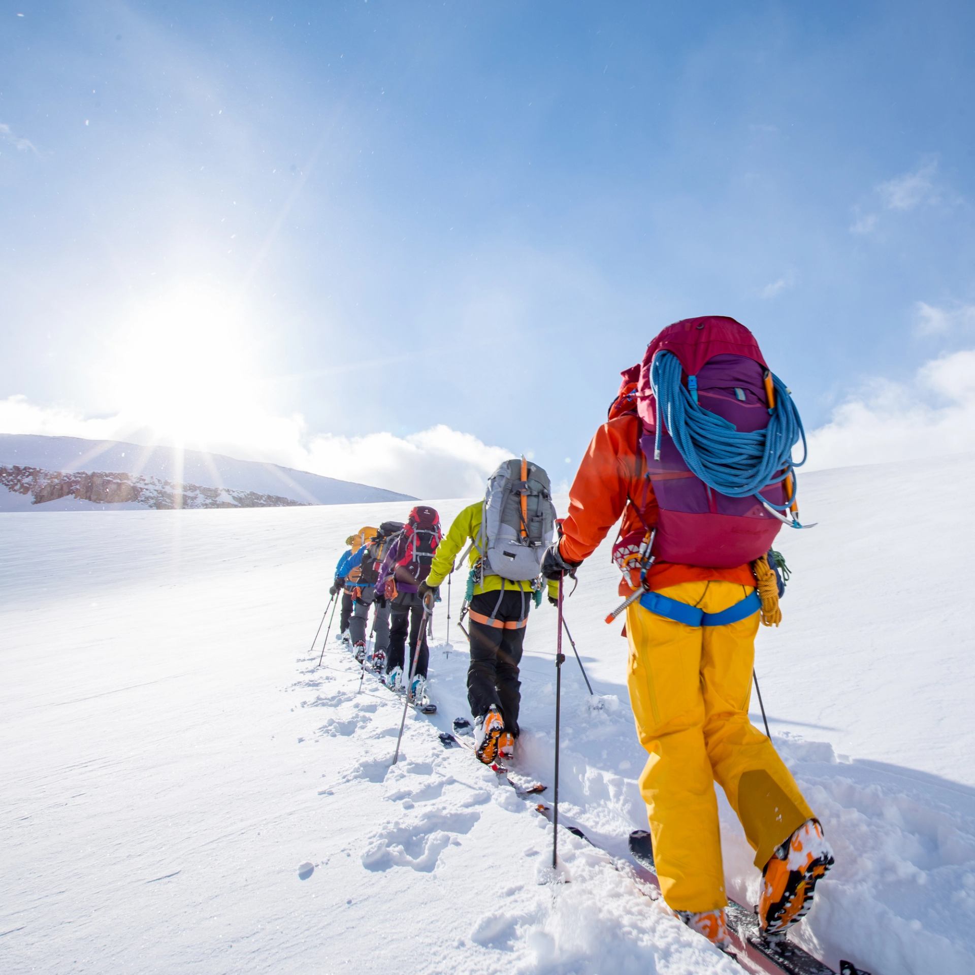 A group of backcountry skiers skinning up a ridge towards the sun that is peaking over the summit.