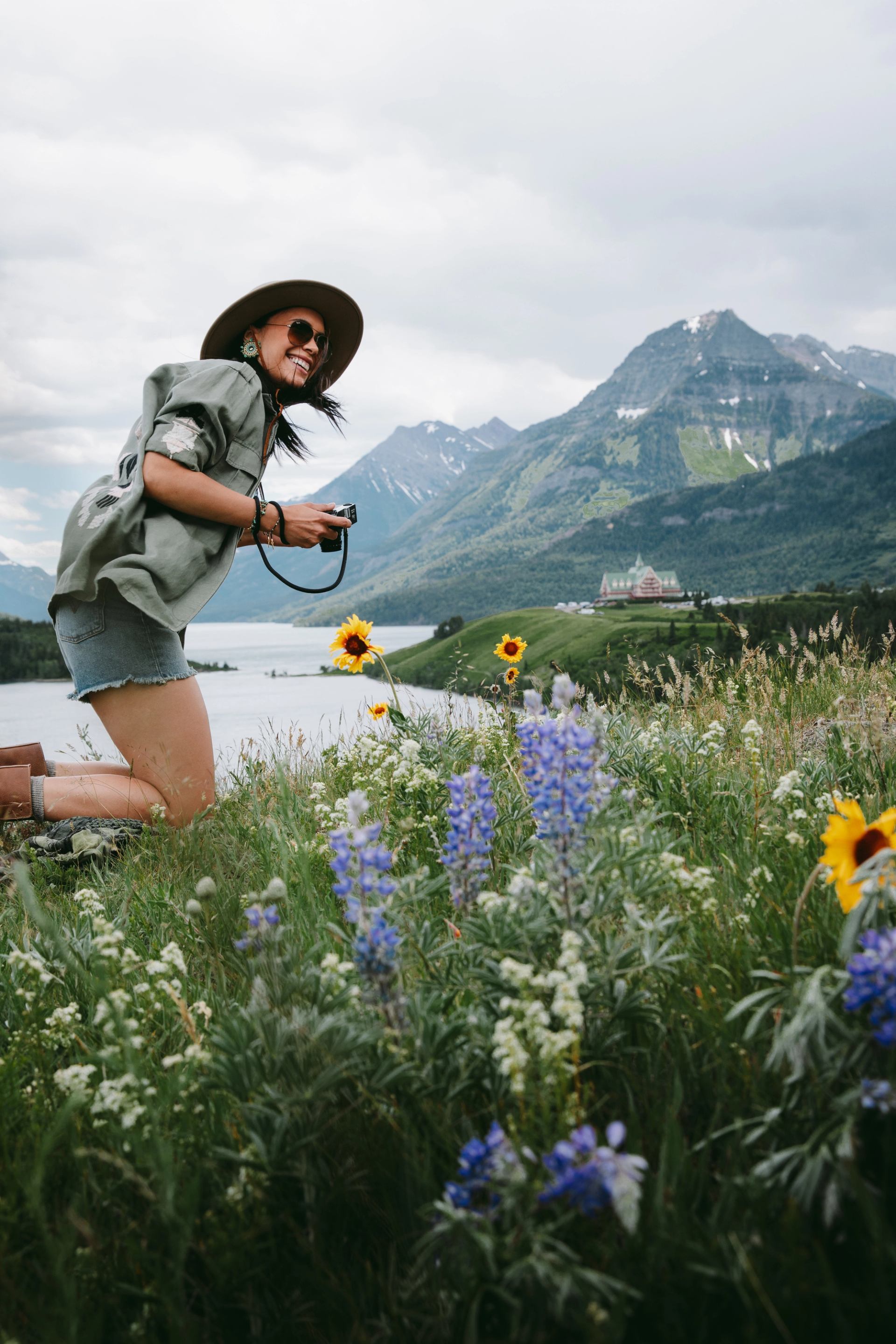 A woman resting on a stone looking at colourful wildflowers, holding a camera with a mountain and lake in the background.
