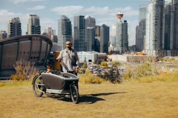 A smiling father stands next to a cargo bike holding his two young kids with Calgary’s downtown skyline in the background.