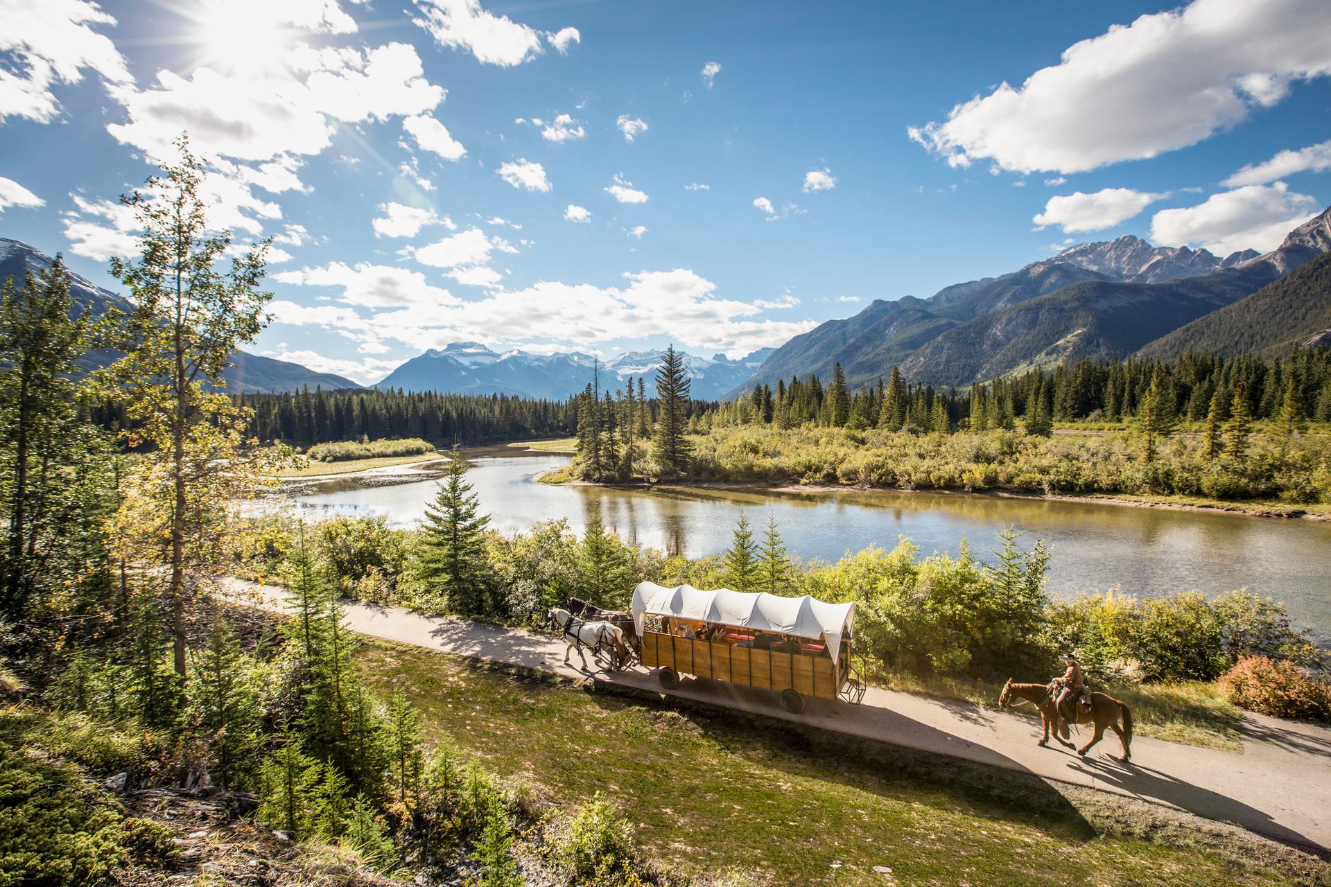 A covered wagon and trail riders travel on a trail along the Bow River in Banff National Park.