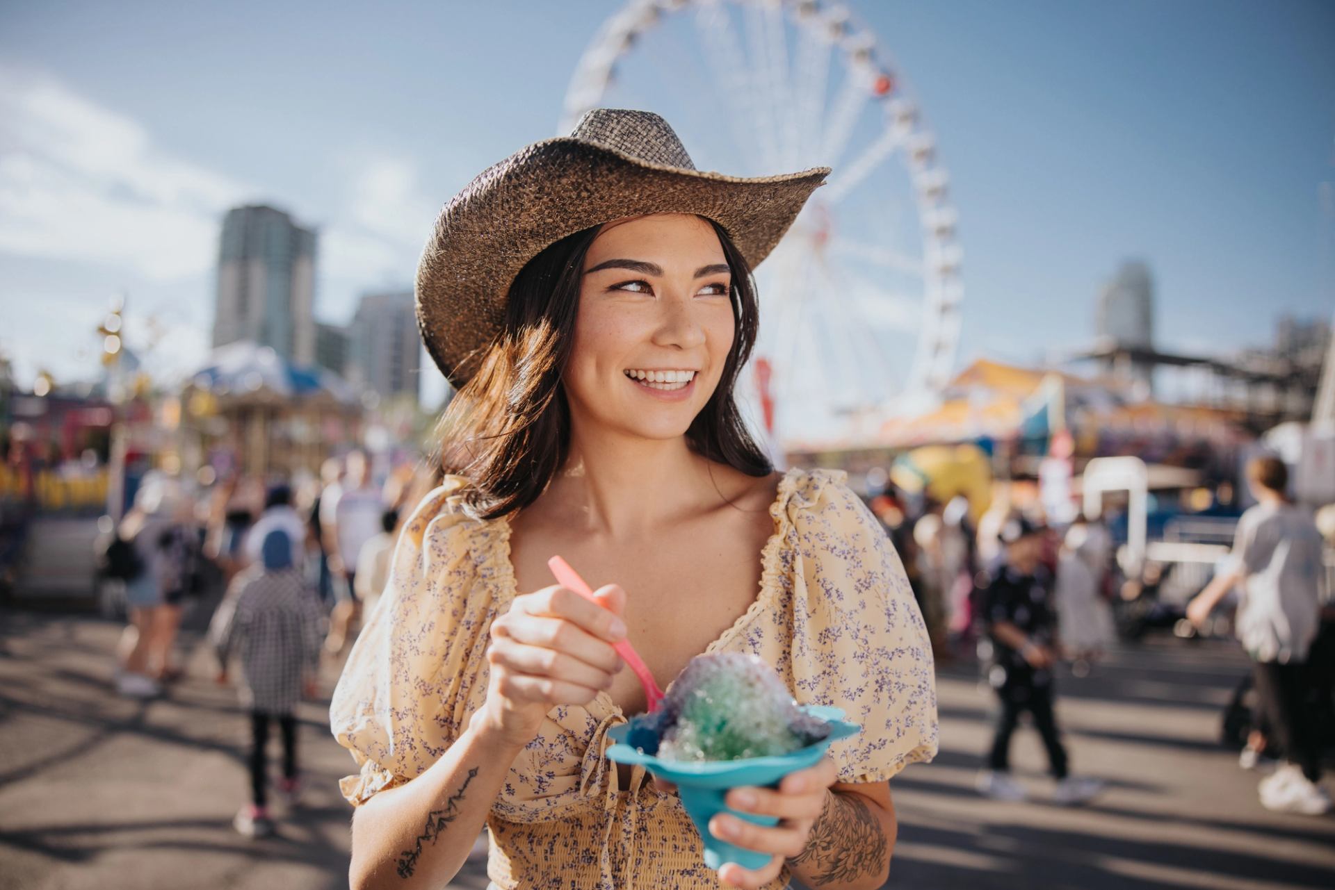 Woman wearing a cowboy hat eats ice cream with midway rides and people in background.