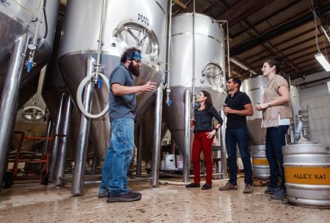 A group of people holding beers take a tour of a brewery.