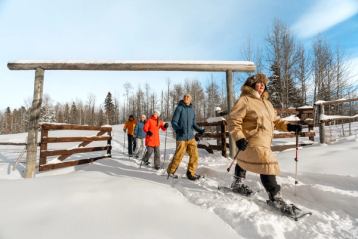 A happy group follow an Indigenous Knowledge Keeper as she guides them on a snowhshoe walk on a sunny day.