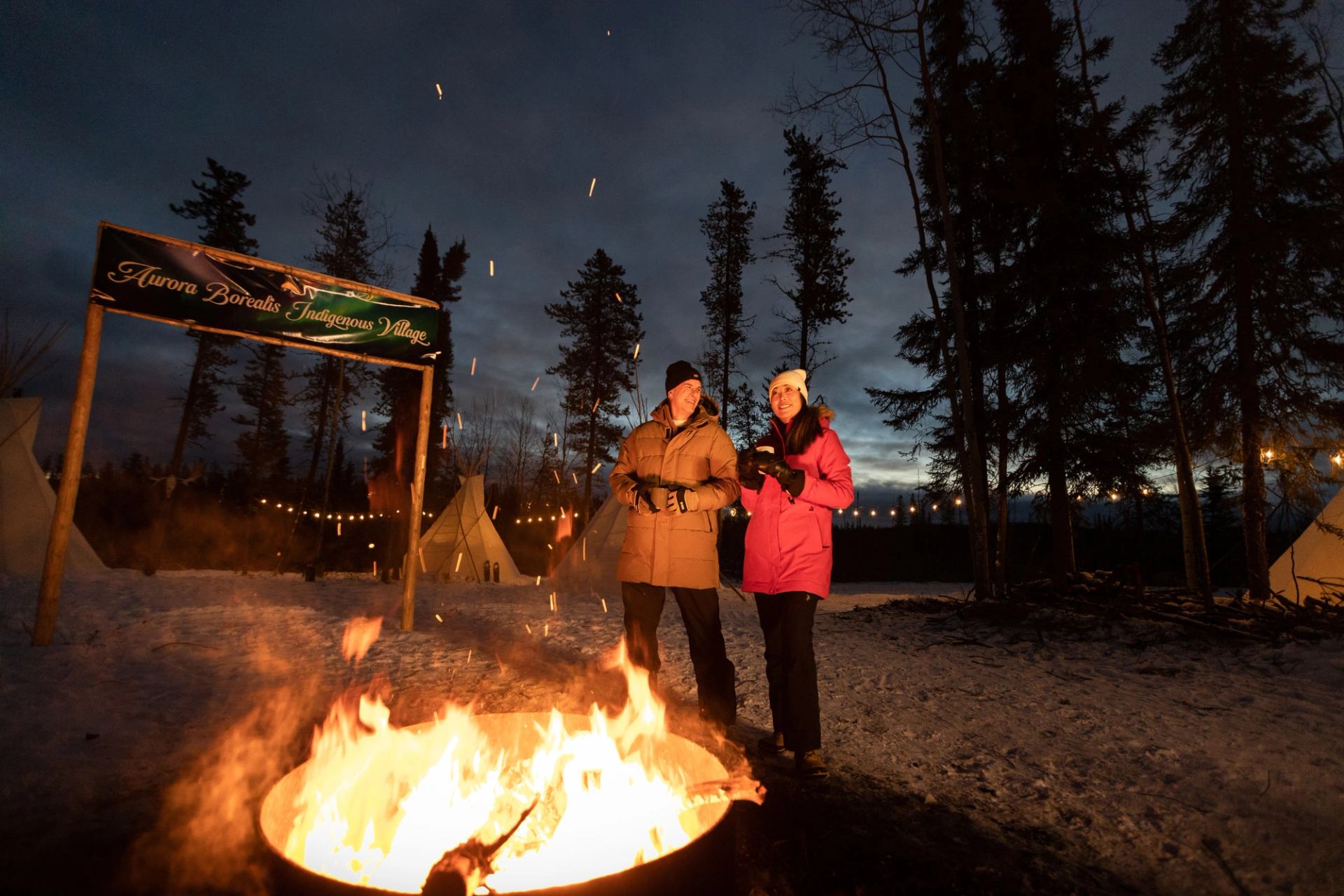 A couple around a fire at night at Aurora Borealis Indigenous Village.