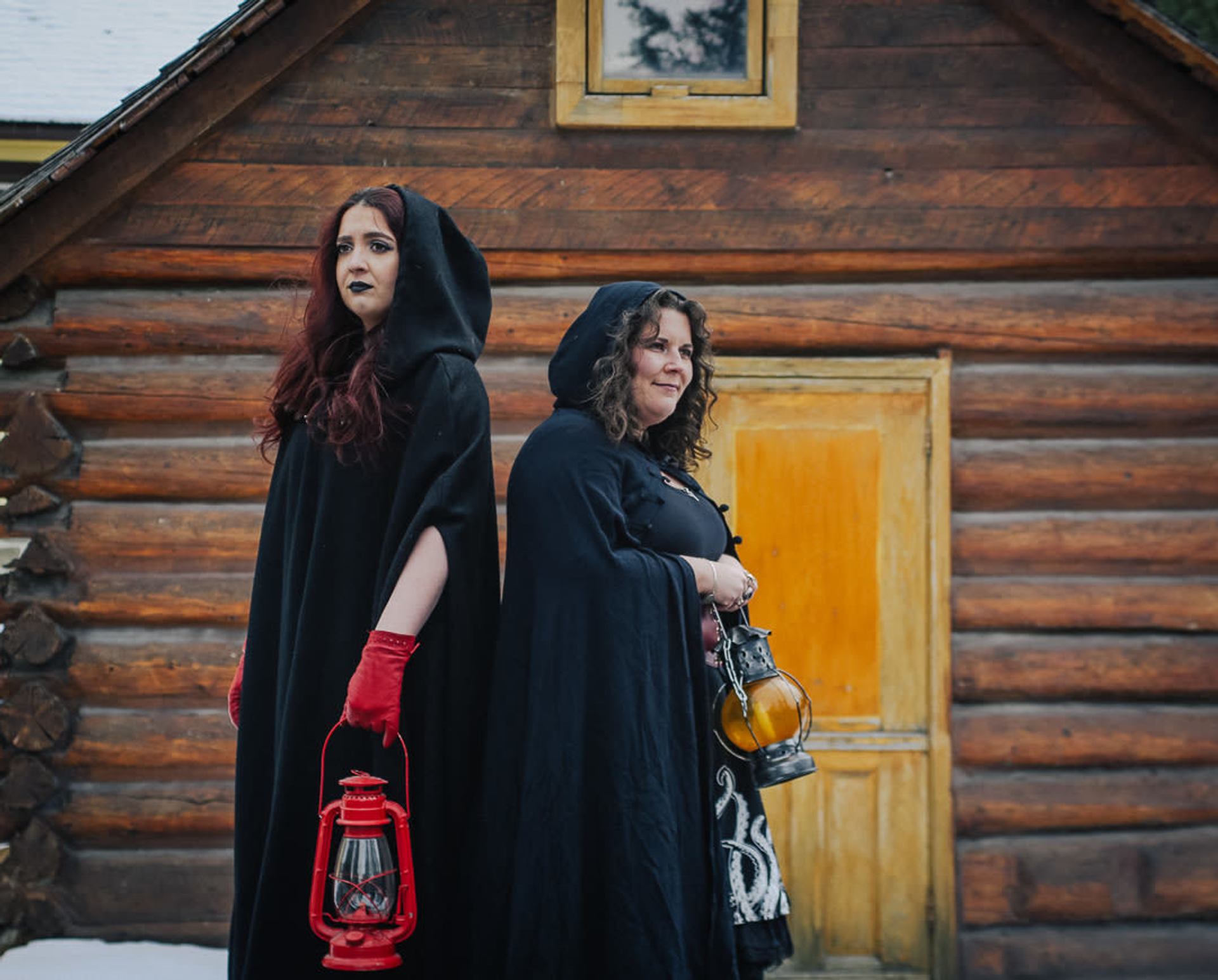 Two women dressed in black Halloween costumes and holding lanterns stand in front of a wooden building.