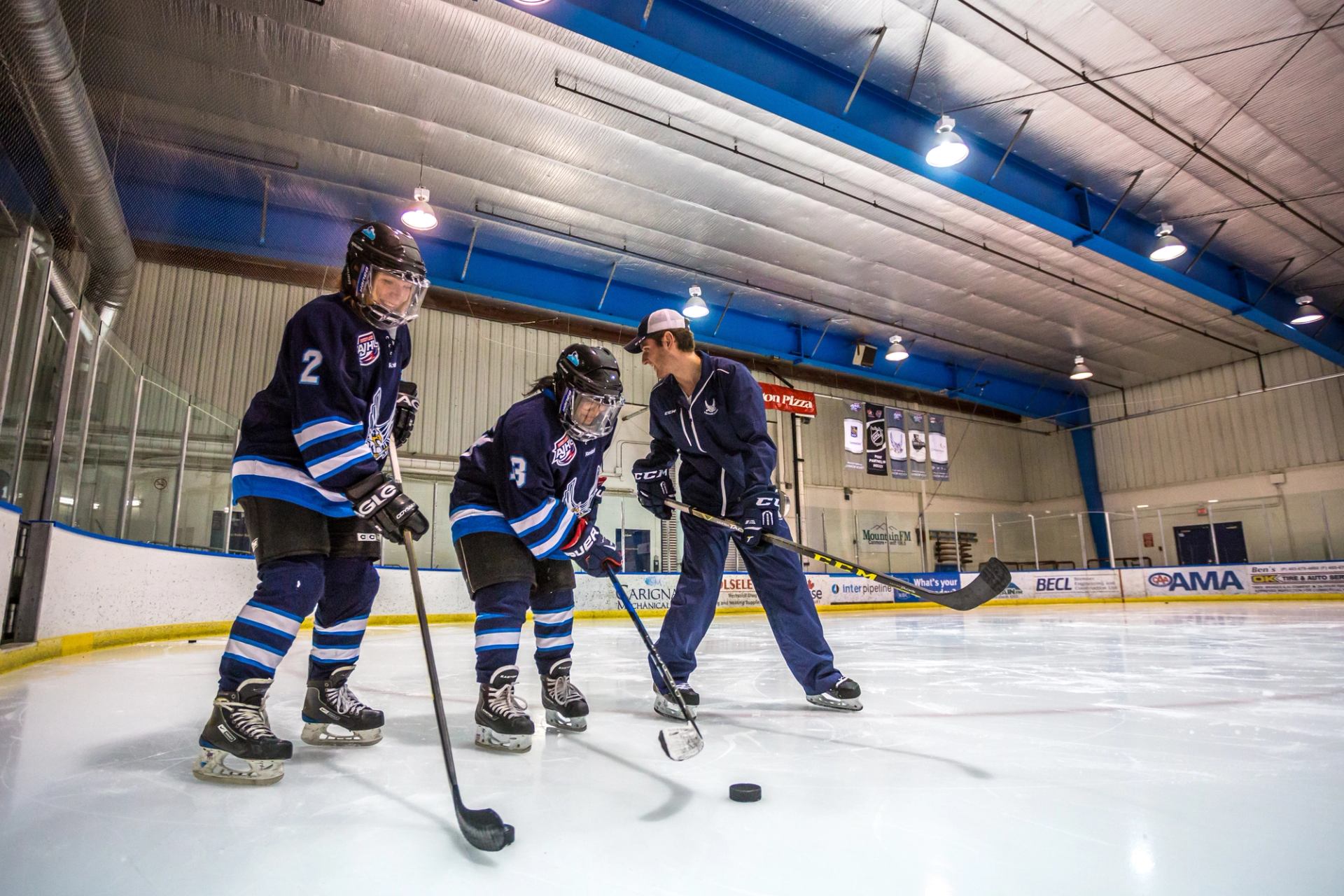Two skaters in full hockey gear work with a coach on hockey rink ice.