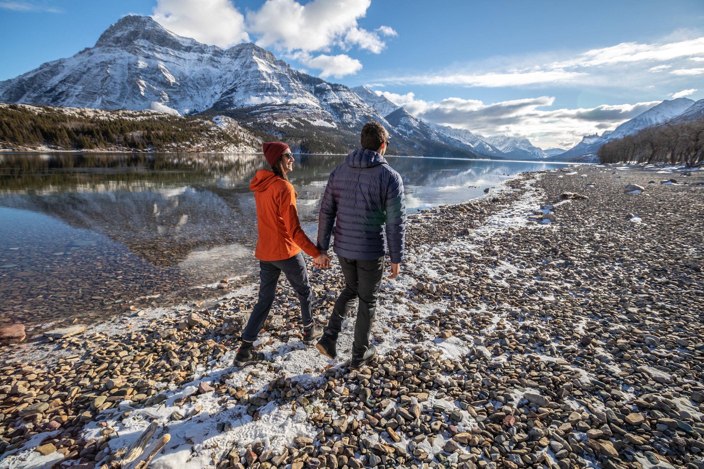 A couple walking the shoreline, mountains in the background, while winter hiking in Waterton Lakes National Park.