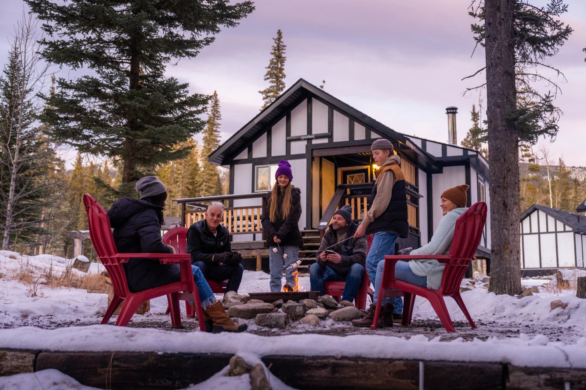 A group gathered around a fire pit outside a rustic cabin surrounded by snow-covered trees.