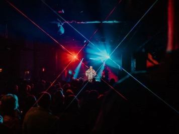 Concert scene with performer on stage under red and blue lights. Audience faces the stage. Light beams form an X pattern in the dark venue.