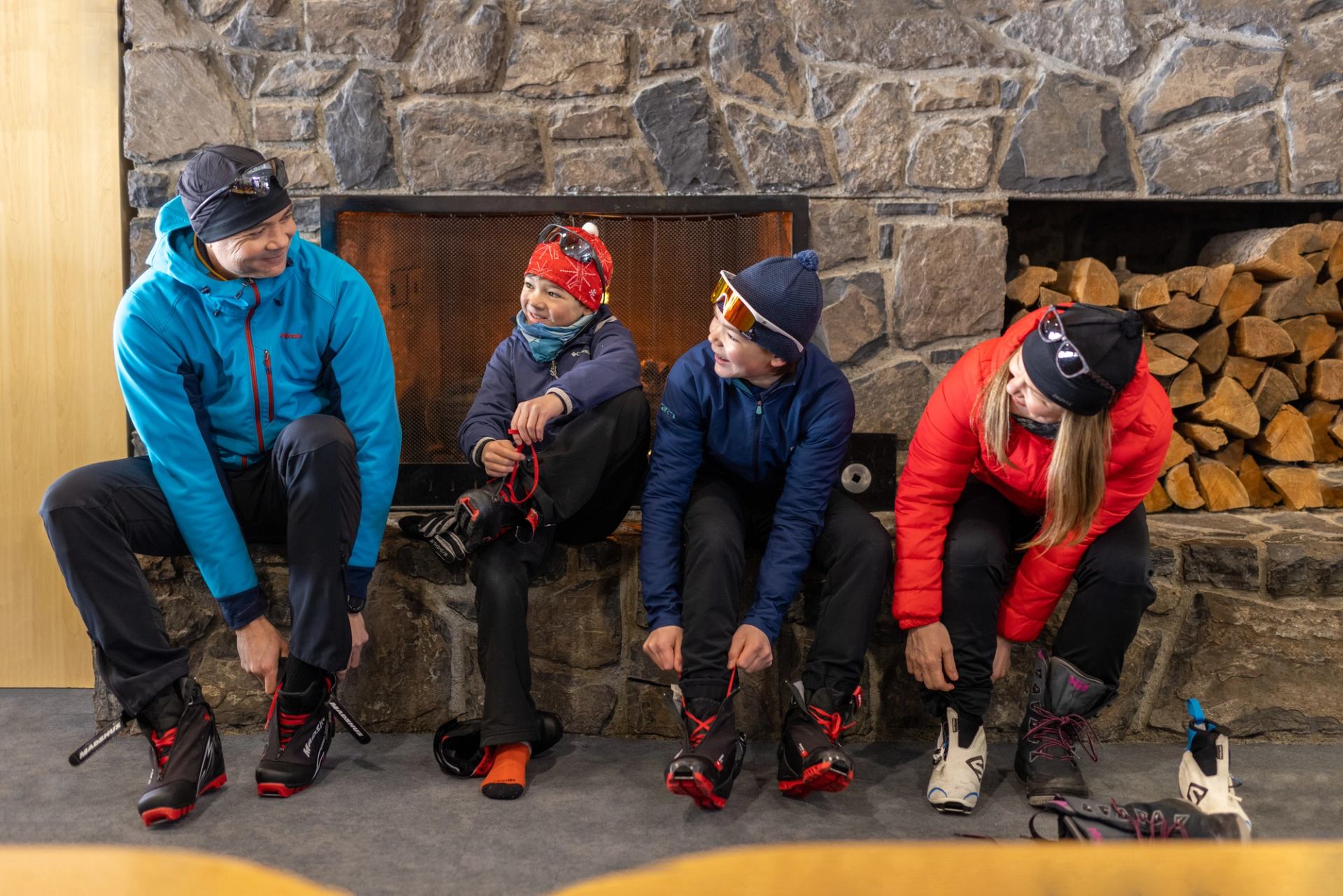 A family of four putting on their ski boots while sitting on a ledge in front of a fireplace.