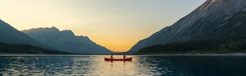 Two people in a red canoe on Spray Lakes, surrounded by the Rocky Mountains at sunset.