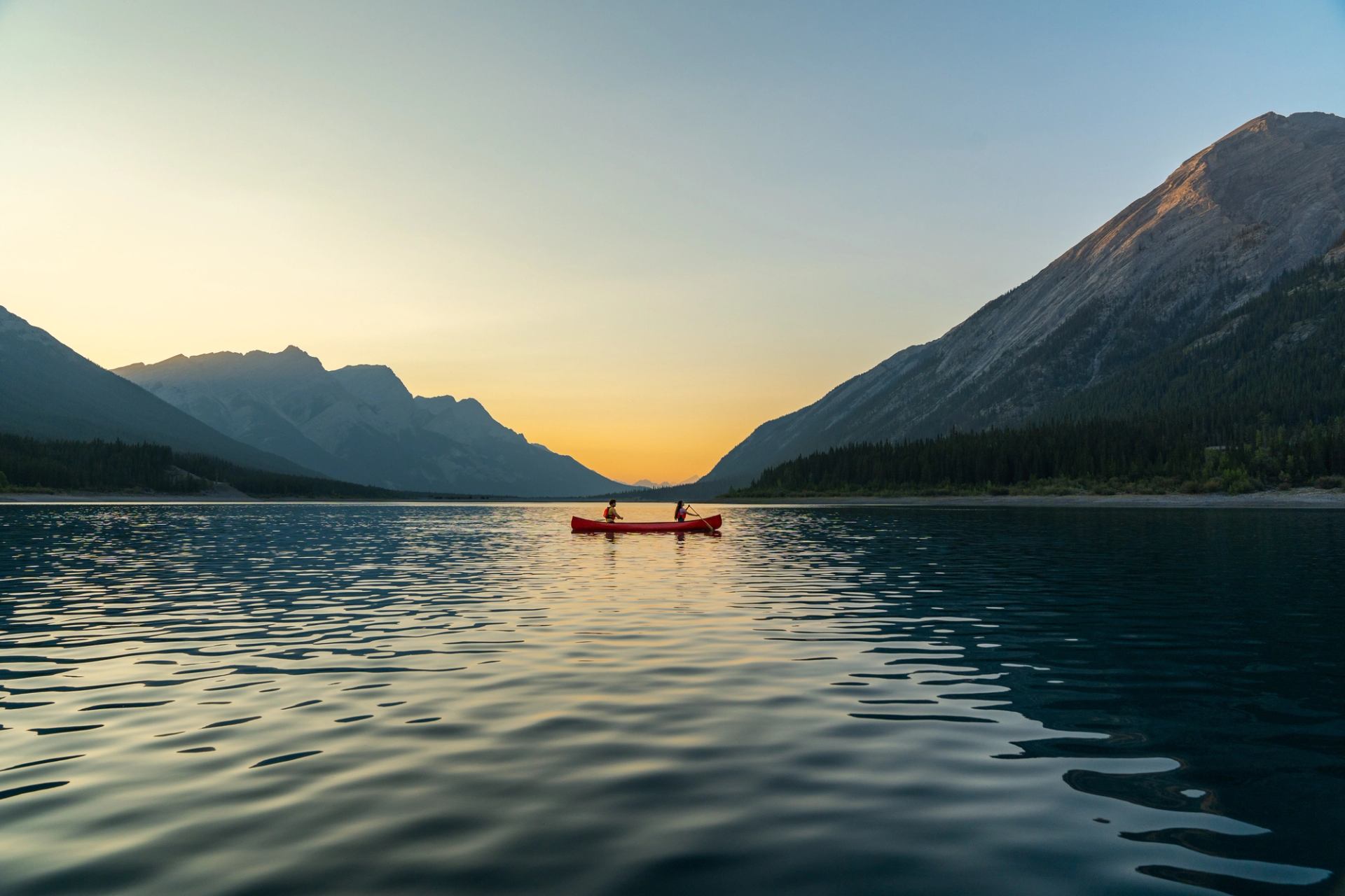 Two people in a red canoe on Spray Lakes, surrounded by the Rocky Mountains at sunset.