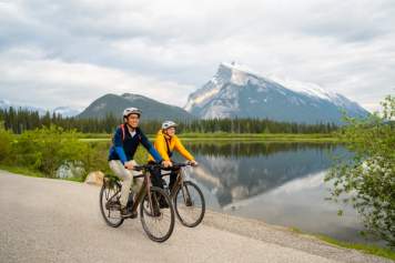 A couple rides bikes on a path with a mountain and lake in the background.