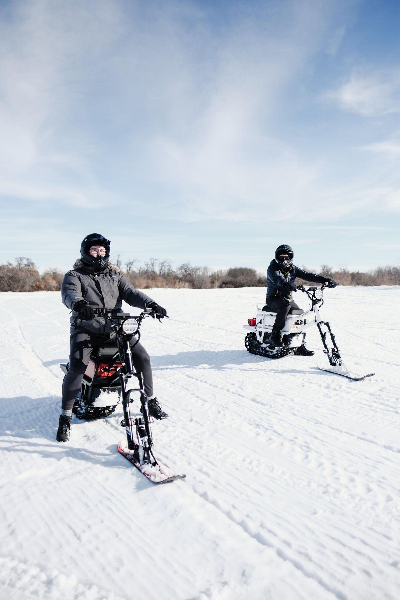 Two warmly dressed people ride MoonBikes, electric bikes with a ski-like front and motorized tread in back. 