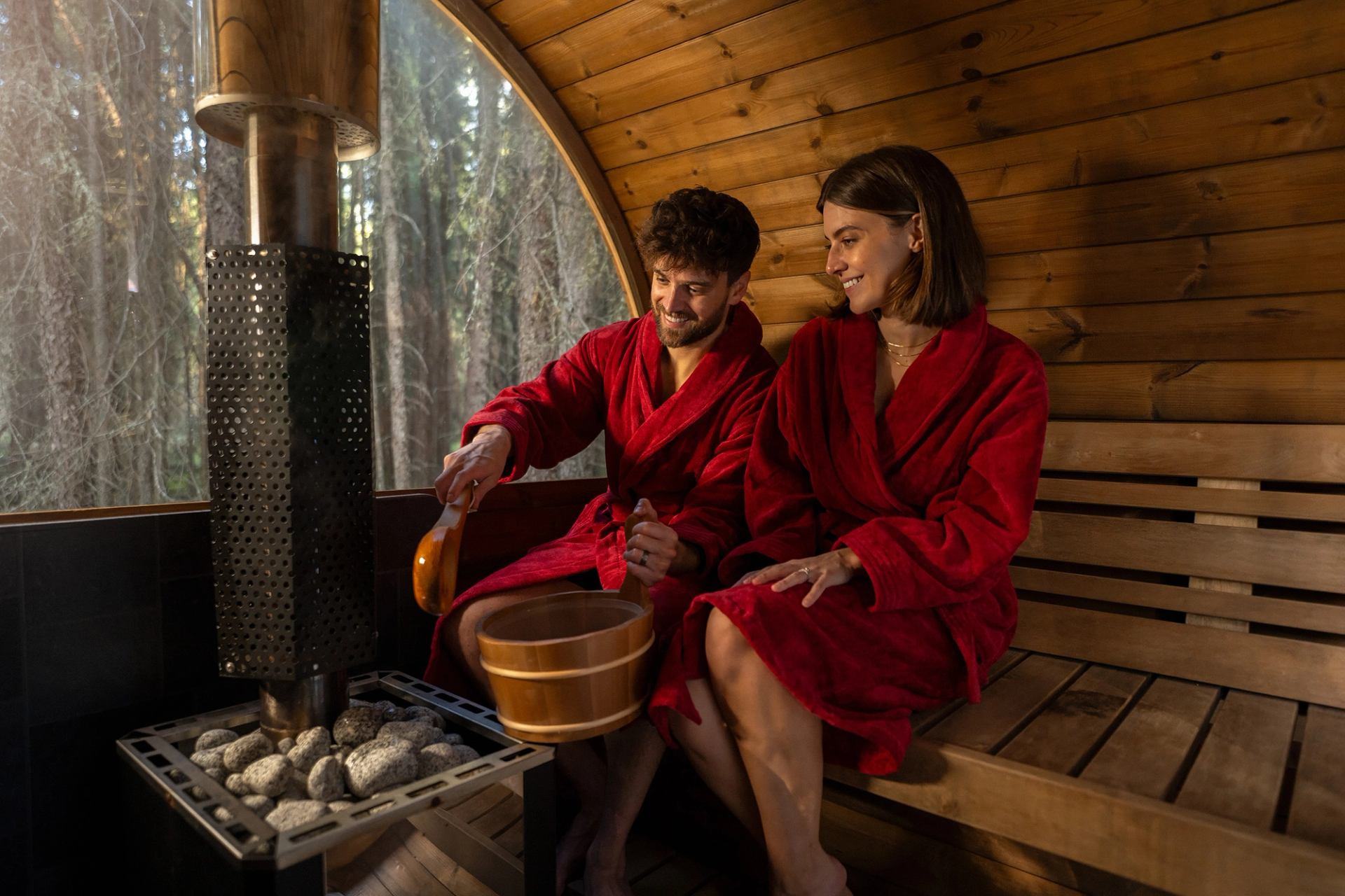 A man and a woman wearing fluffy red robes sit inside a barrel sauna, pouring water on hot rocks.