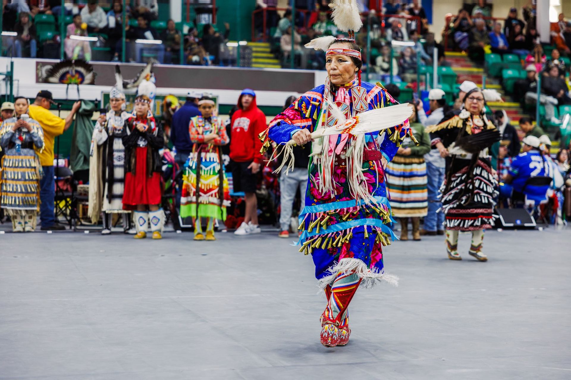 A female dancer holding a feather fan and wearing a blue jingle dress trimmed with pink and cream details and metal cones.