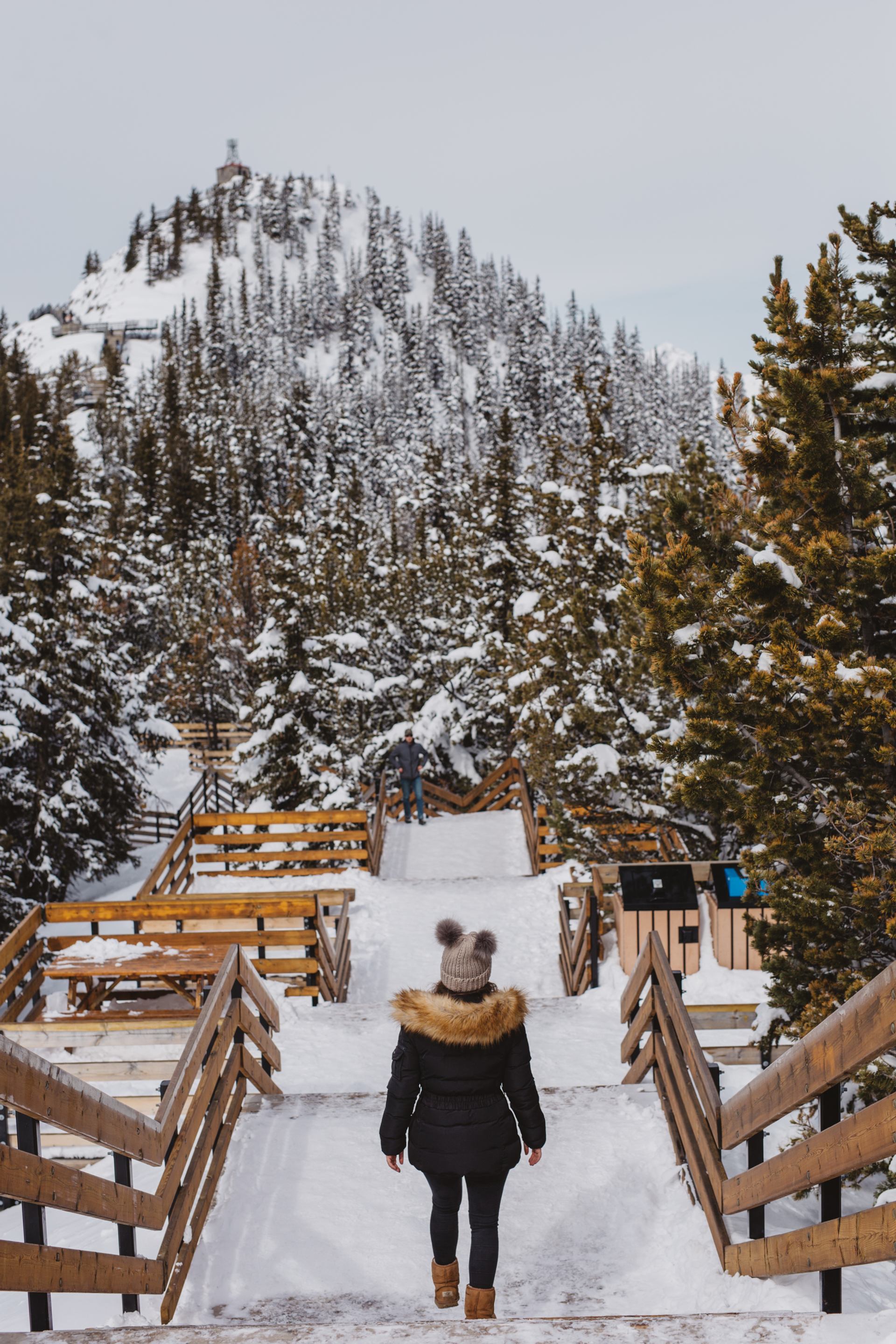 A visitor in a fur-lined parka walks the snowy boardwalk at the top of the Banff Gondola in Banff National Park.