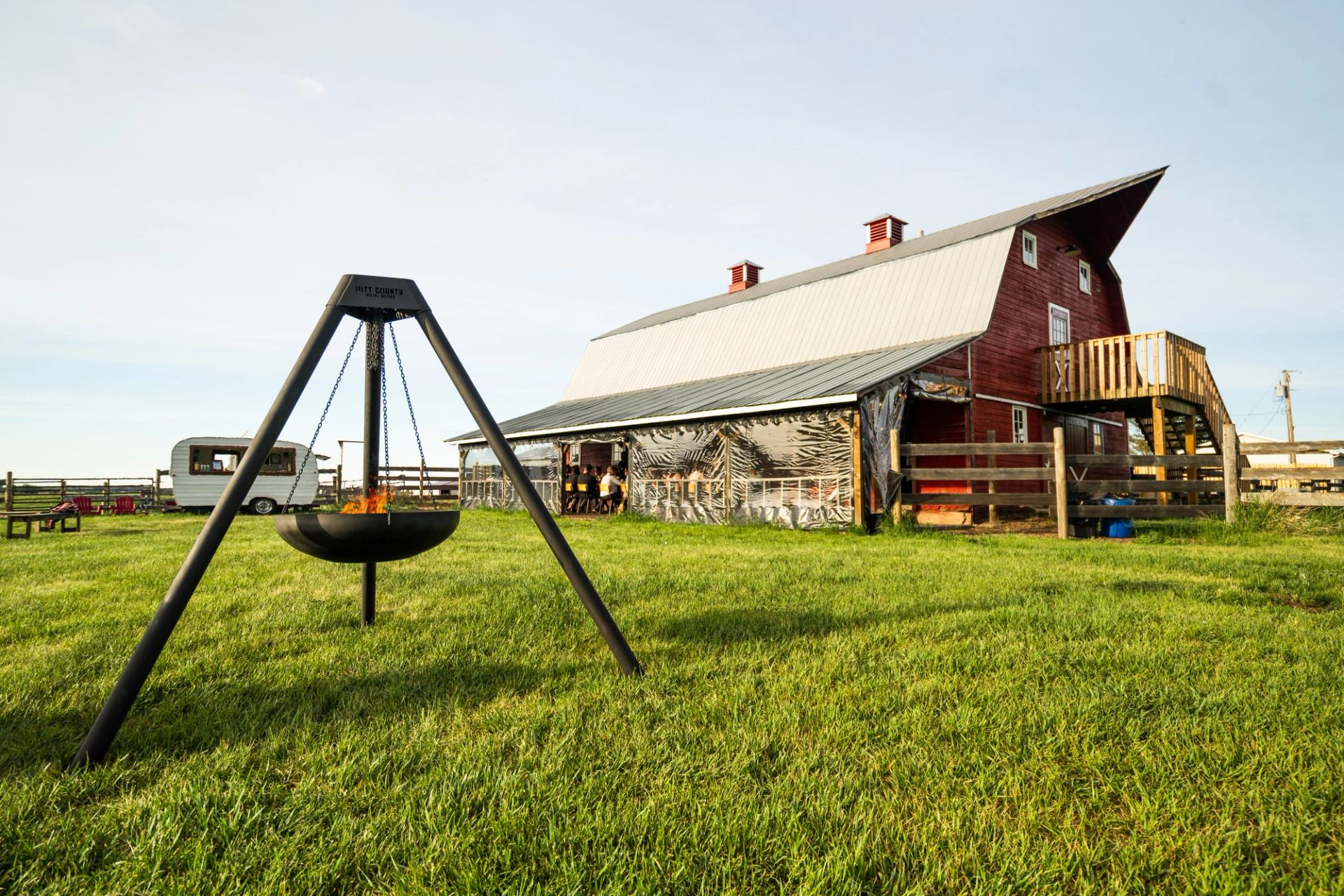 A bird's-eye view of an old red barn amidst greenery. A wooden table is set outdoors with dinnerware, glasses, and candles. Flames rise from a nearby grill, enhancing the cozy, rustic atmosphere, inviting guests to a farm-to-table dining experience