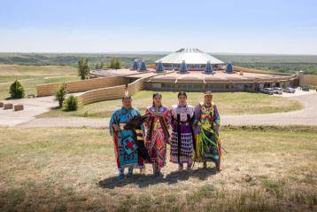 Four women in traditional Indigenous clothing stand in front of the visitor centre at Blackfoot Crossing Historical Park.