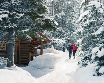 Couple walking through the snow covered trees to a log cabin at Storm Mountain Lodge in Banff National Park