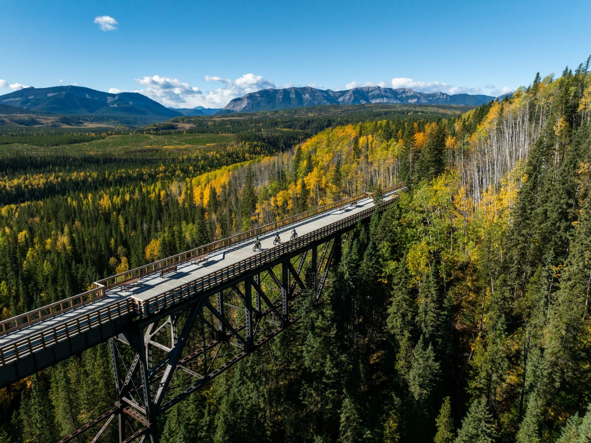 Cyclists riding across a wooden trestle bridge high above an autumn forest and surrounded by mountain peaks.