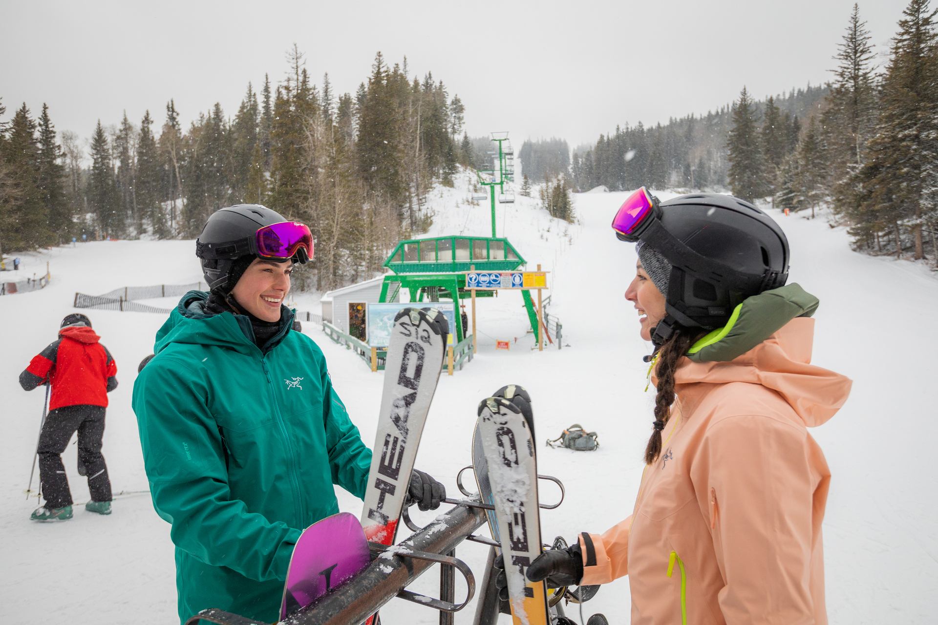 Two skiers retrieve their skis from a rack at the base of the hill with a chairlift in the background.