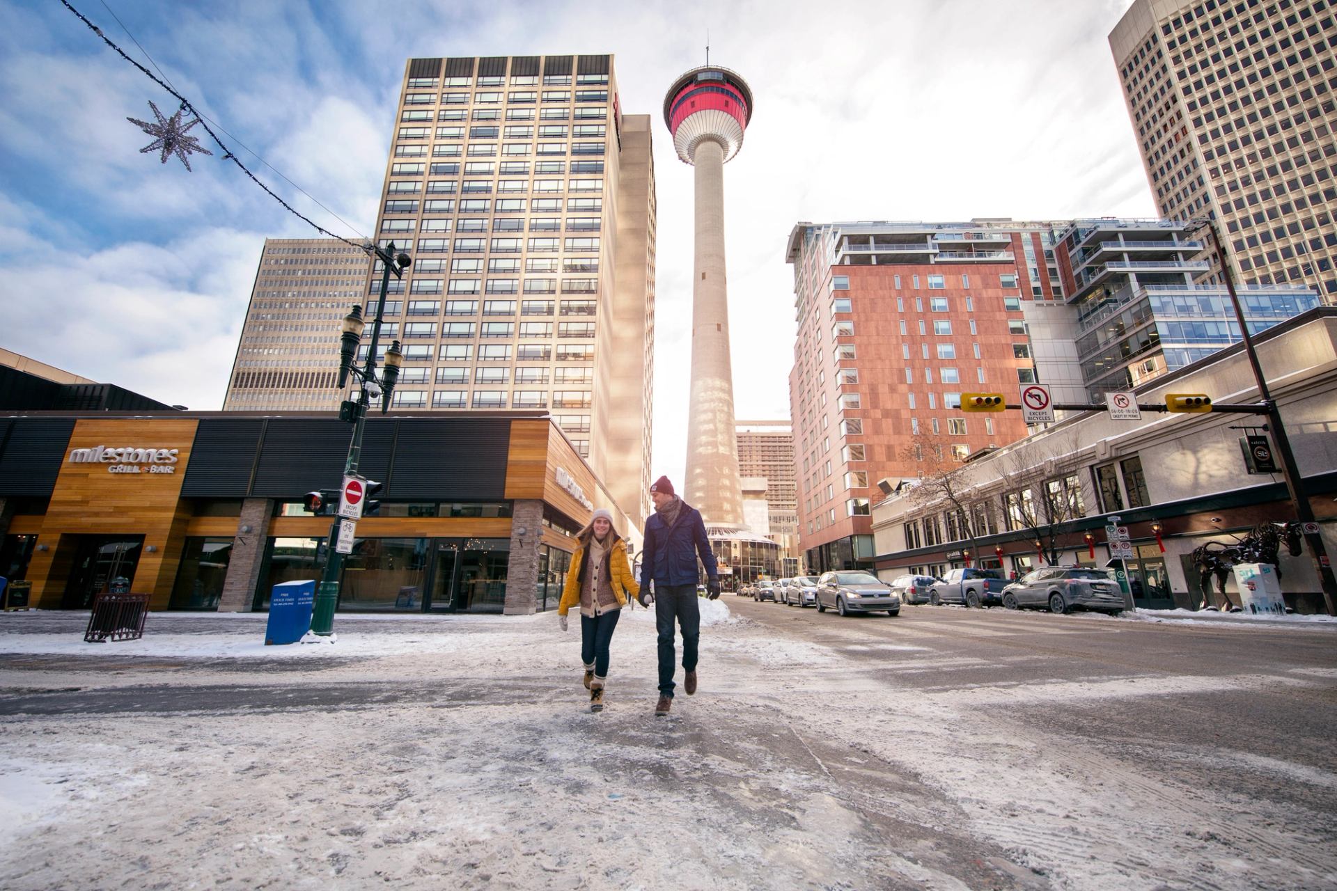 Couple walking down a snowy street in the winter, with the Calgary Tower and downtown buildings in the background