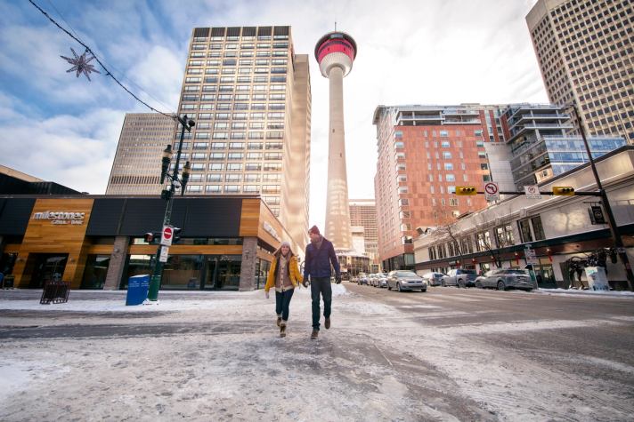 Couple walking down a snowy street in the winter, with the Calgary Tower and downtown buildings in the background