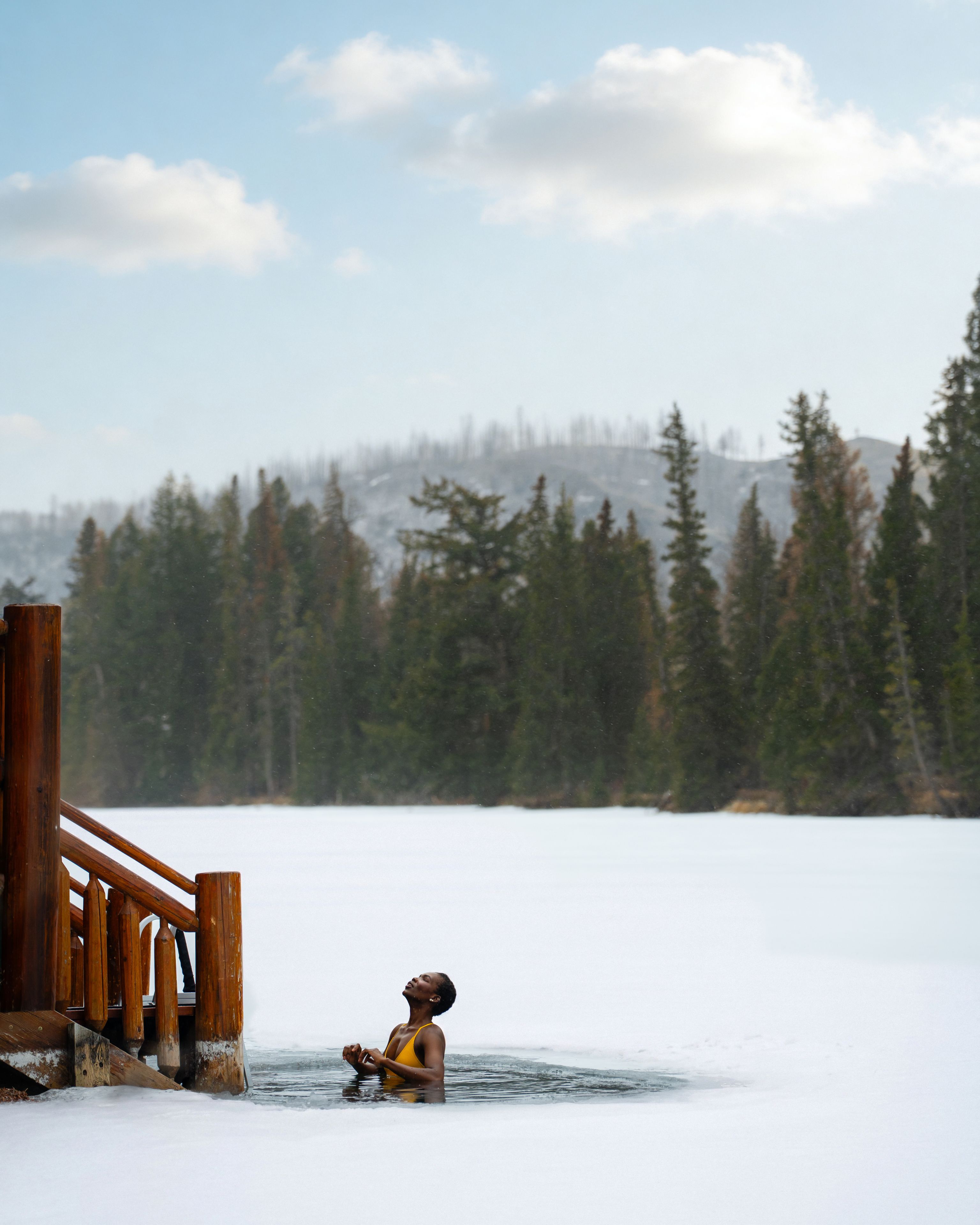 A guest cold-plunges in a hole in the ice off the shore Lac Beauvert with trees in the distance. 