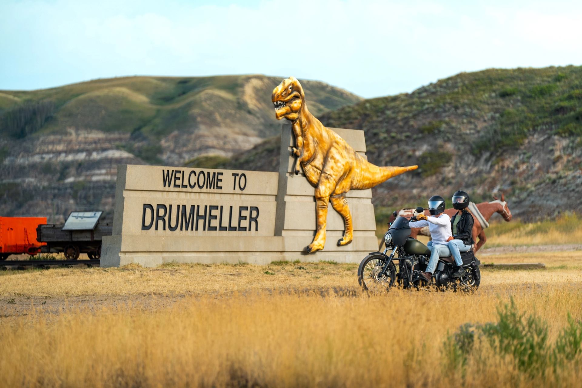 Two people riding a motorcycle pass the dinosaur on the “Welcome to Drumheller” sign.