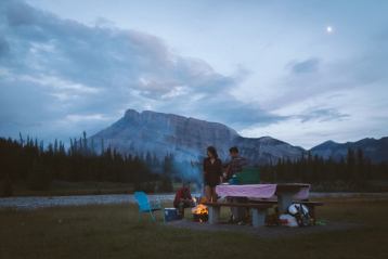 Friends around a campfire next to a picnic bench with a mountain view behind