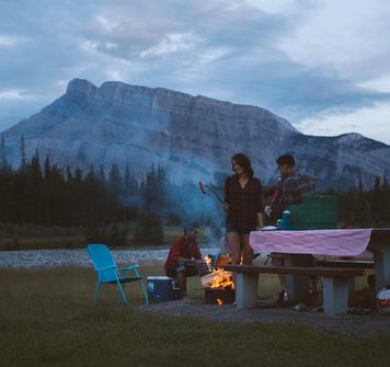 Friends around a campfire next to a picnic bench with a mountain view behind