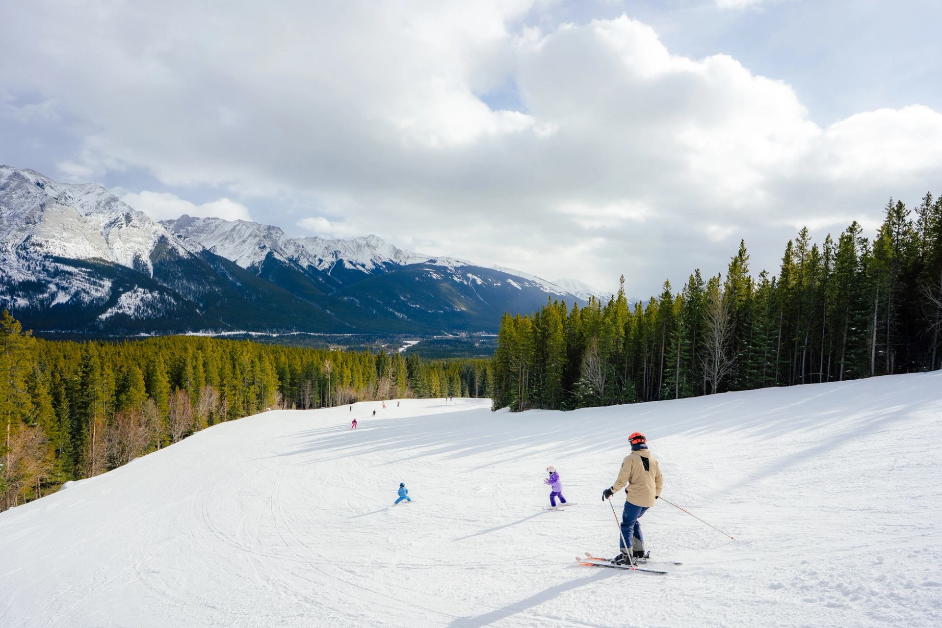 A family of skiers, including two small children, ski down an open trail at Nakiska Ski Resort with mountain views in the distance.
