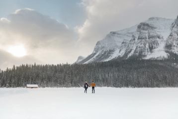 A couple holding hands while ice skating on Lake Louise, mountains and trees in the background.