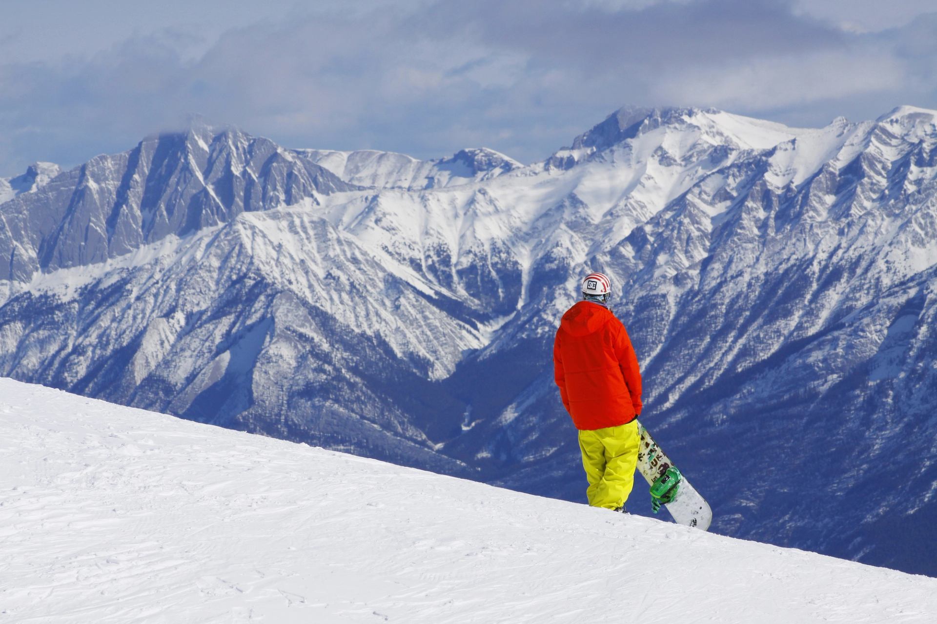 A snowboarder holding their board while looking at the mountain view from the top of a ski resort run.