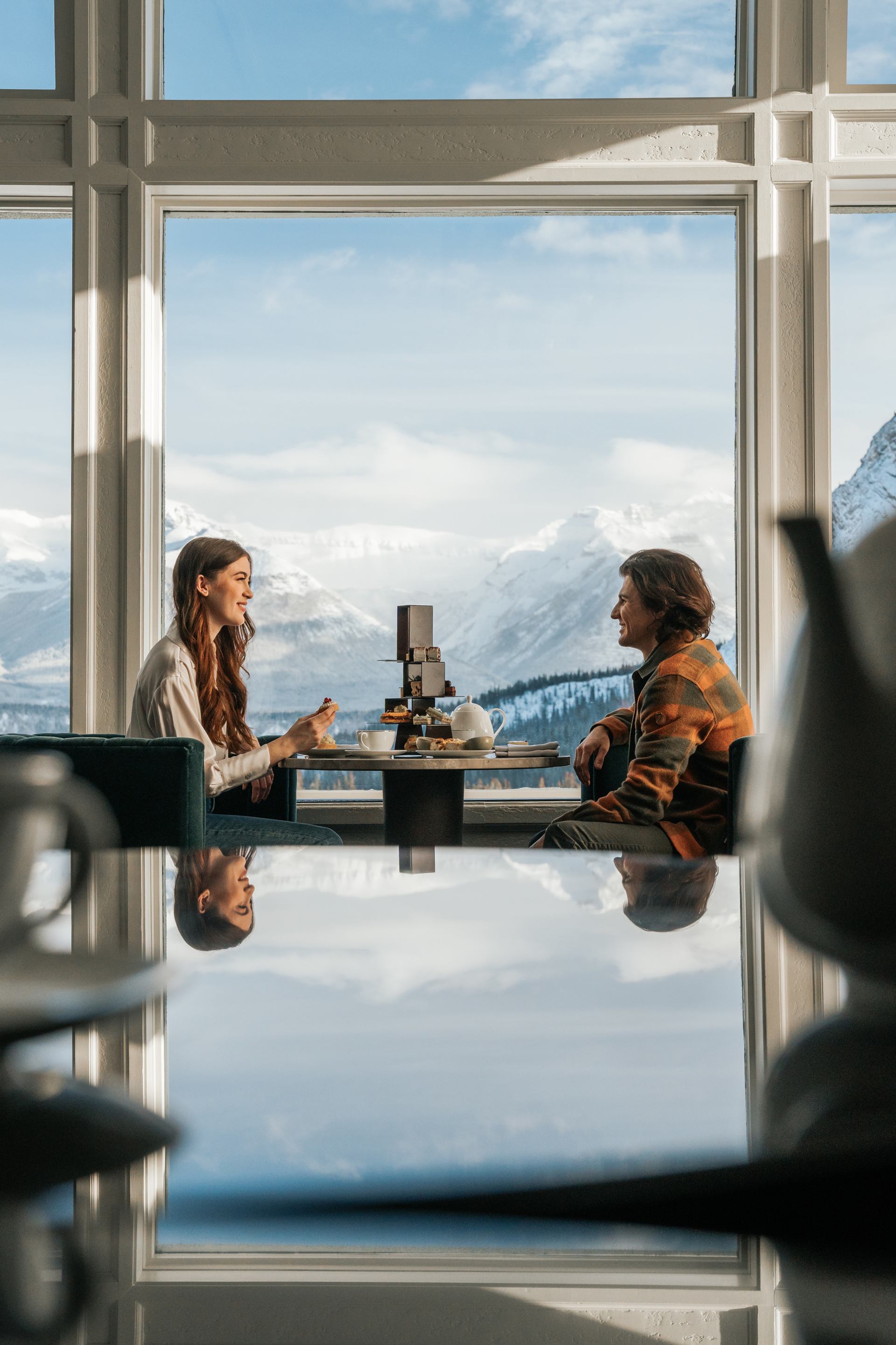 A couple sitting indoors and smiling at each other as they share tea and dessert.