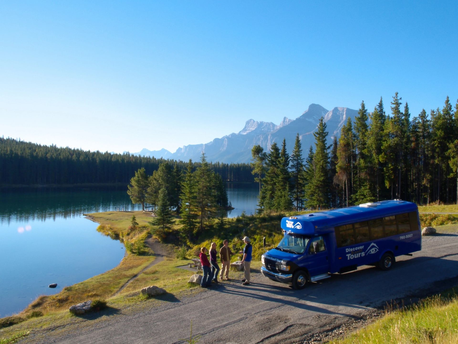 The Discover Banff tour bus stops in front of an alpine lake with a tour group during a wildlife tour.