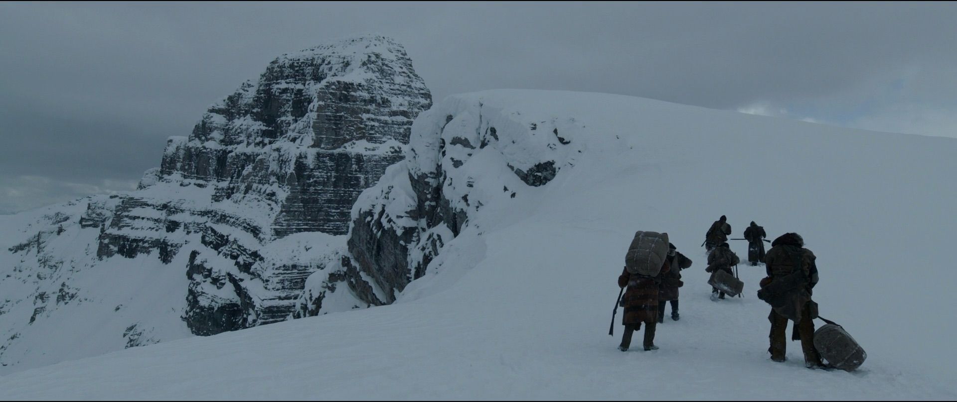 A scene from the Revenant, with characters trudging through snow near a mountain summit on a grey day.