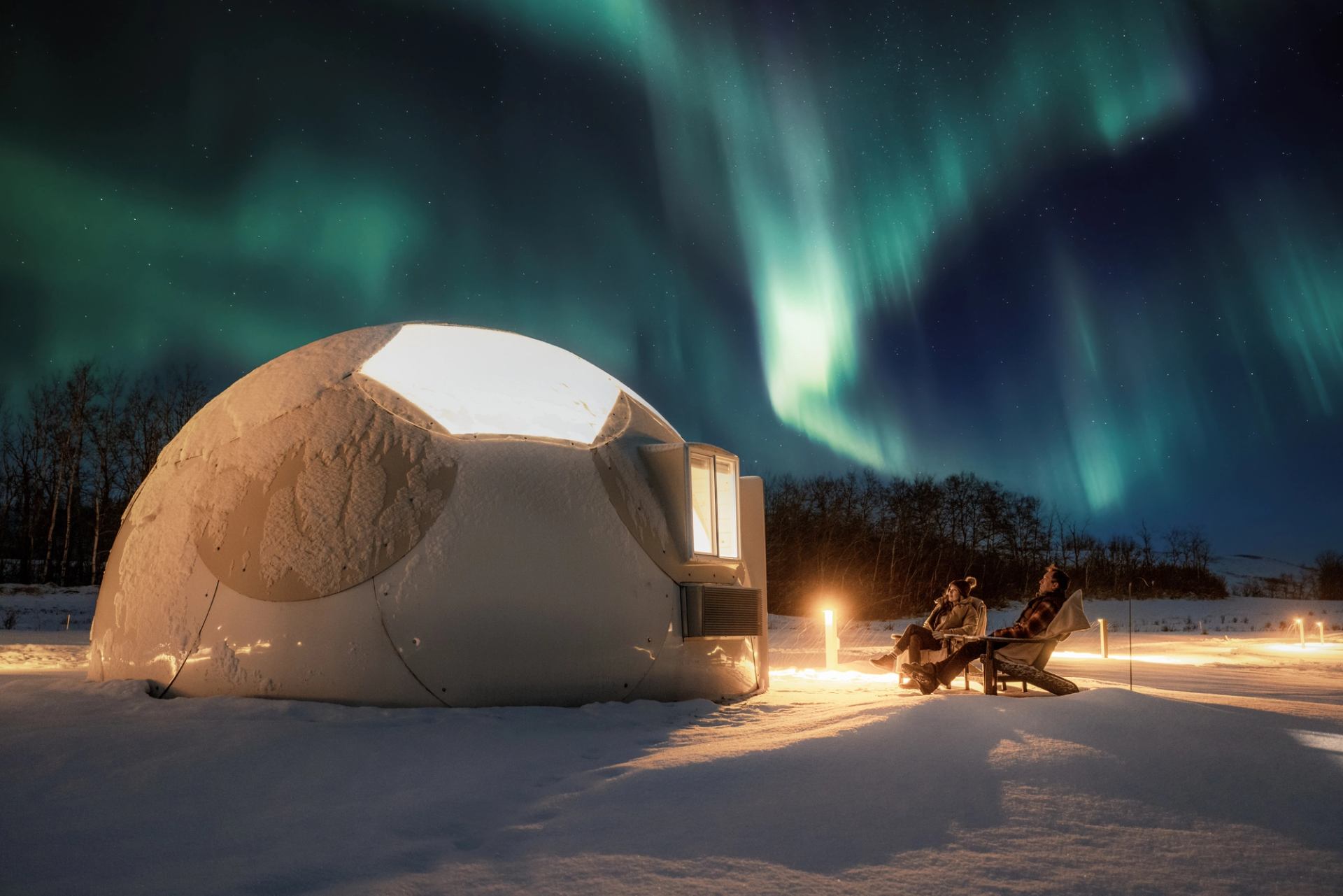 Two people relax outside a geodome while green northern light sparkle above.