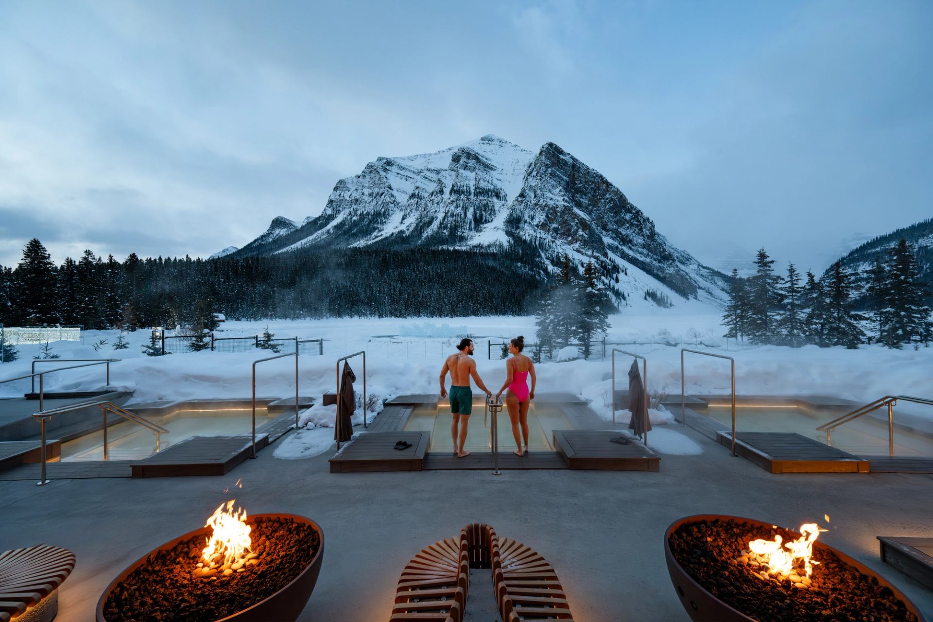 A couple walks into one of 3 luxurious outdoor hot tubs with a snowy mountain in the distance.