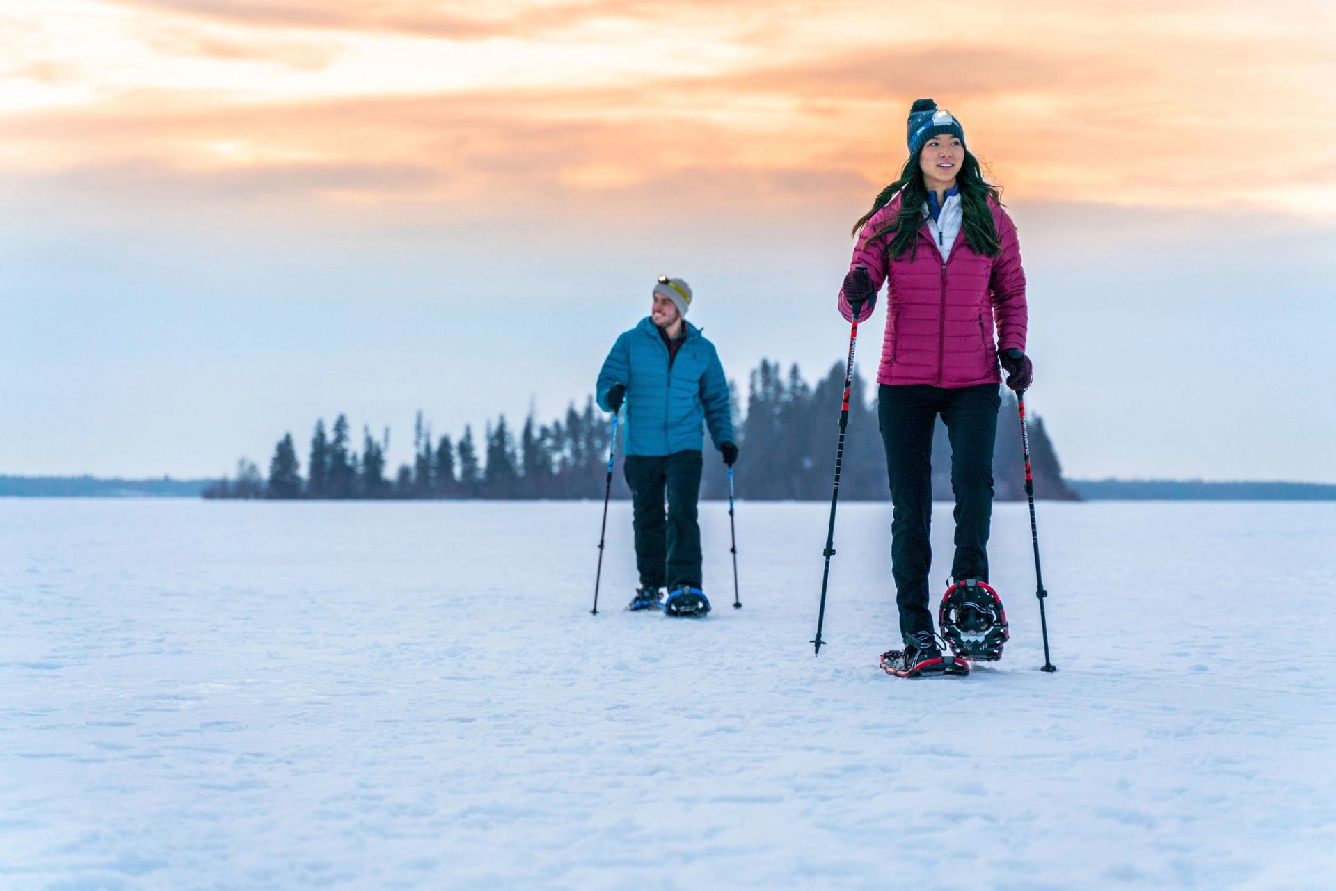 A women with a man following behind, both smiling and walking over snow packed ground on snowshoes.