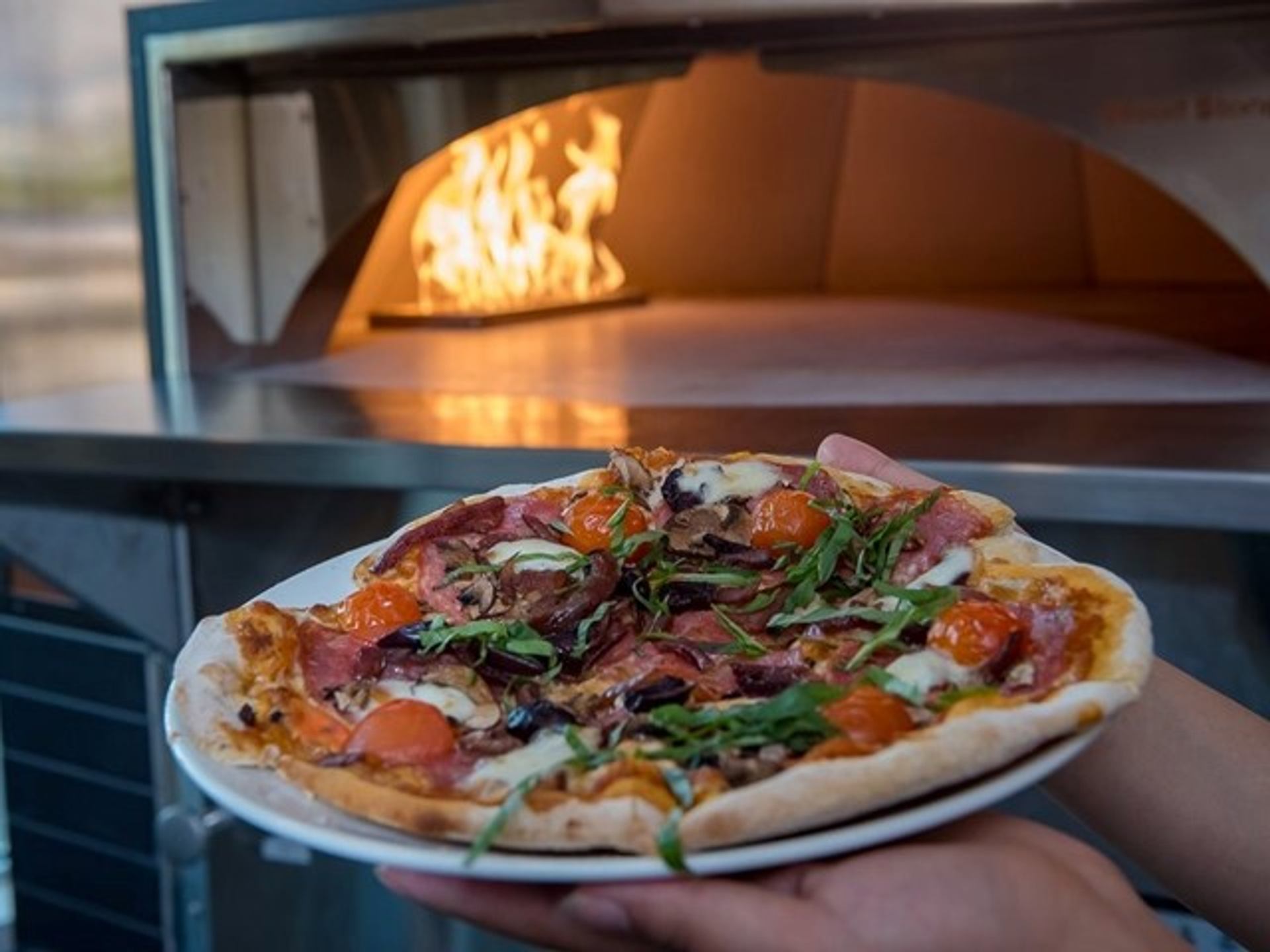 A person holds a pizza in front of a pizza oven.