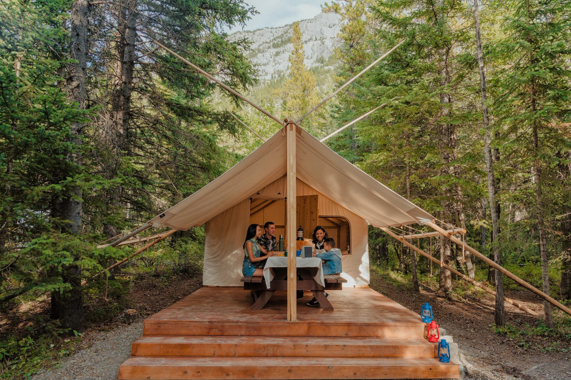 A family sits at a covered picnic table outside their glamping tent in the Rockies, surrounded by forest.