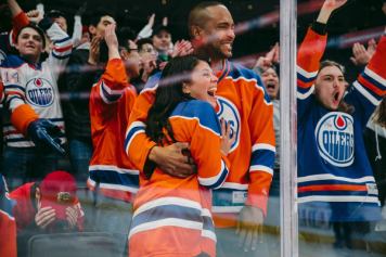 Edmonton Oilers fans cheer behind the glass at a hockey game. One Calgary Flames fan in red jersey and hat sits with head in hands. Others hug and celebrate.