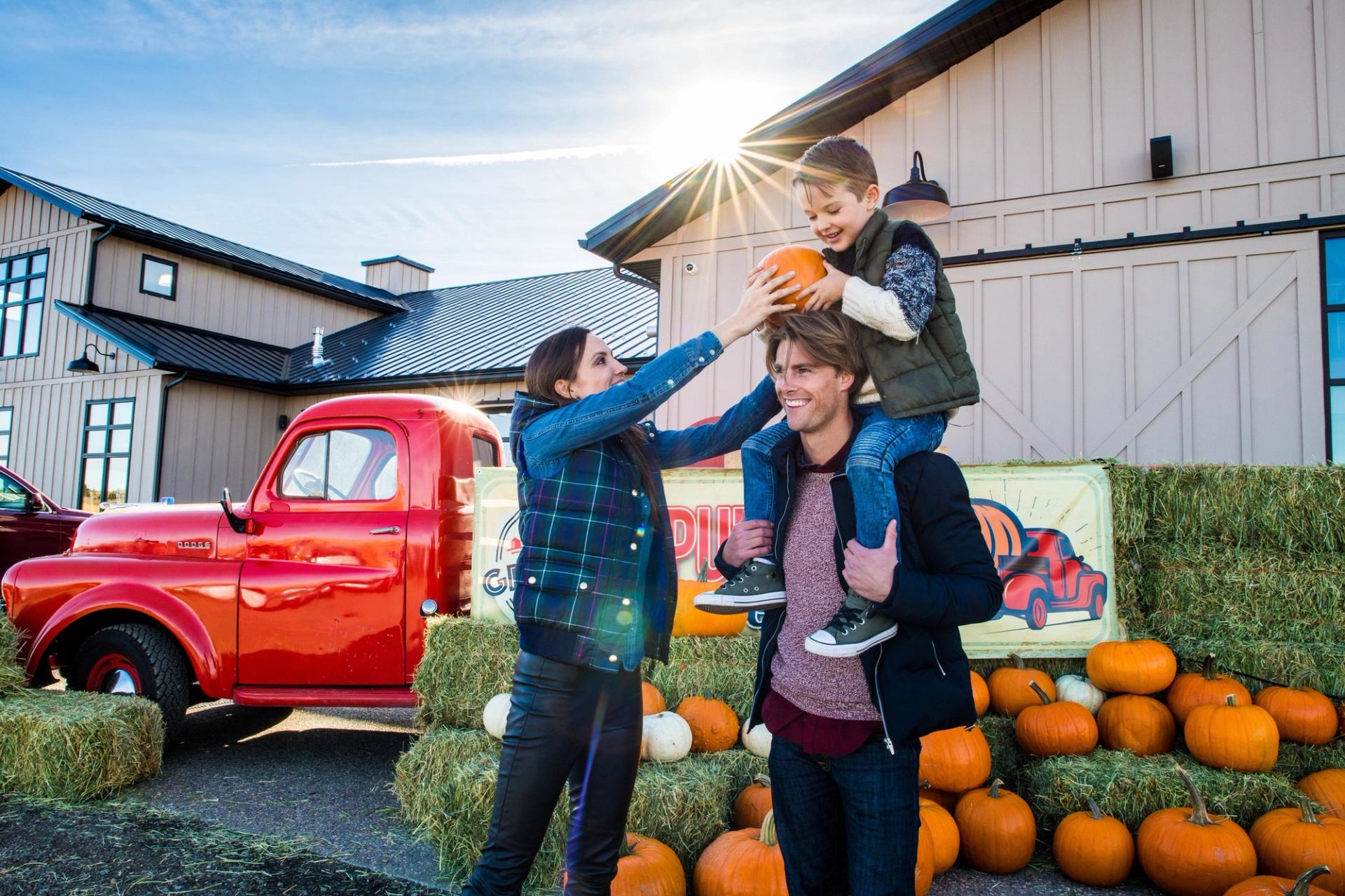 Son on his fathers shoulders as the mom passes him a pumpkin, all smiling as they enjoy an outdoor farmers market.