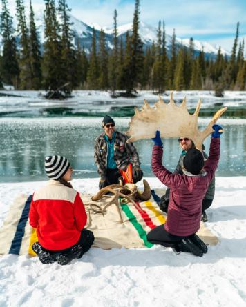 People look at antlers on a blanket while sitting by a snowy riverbank
