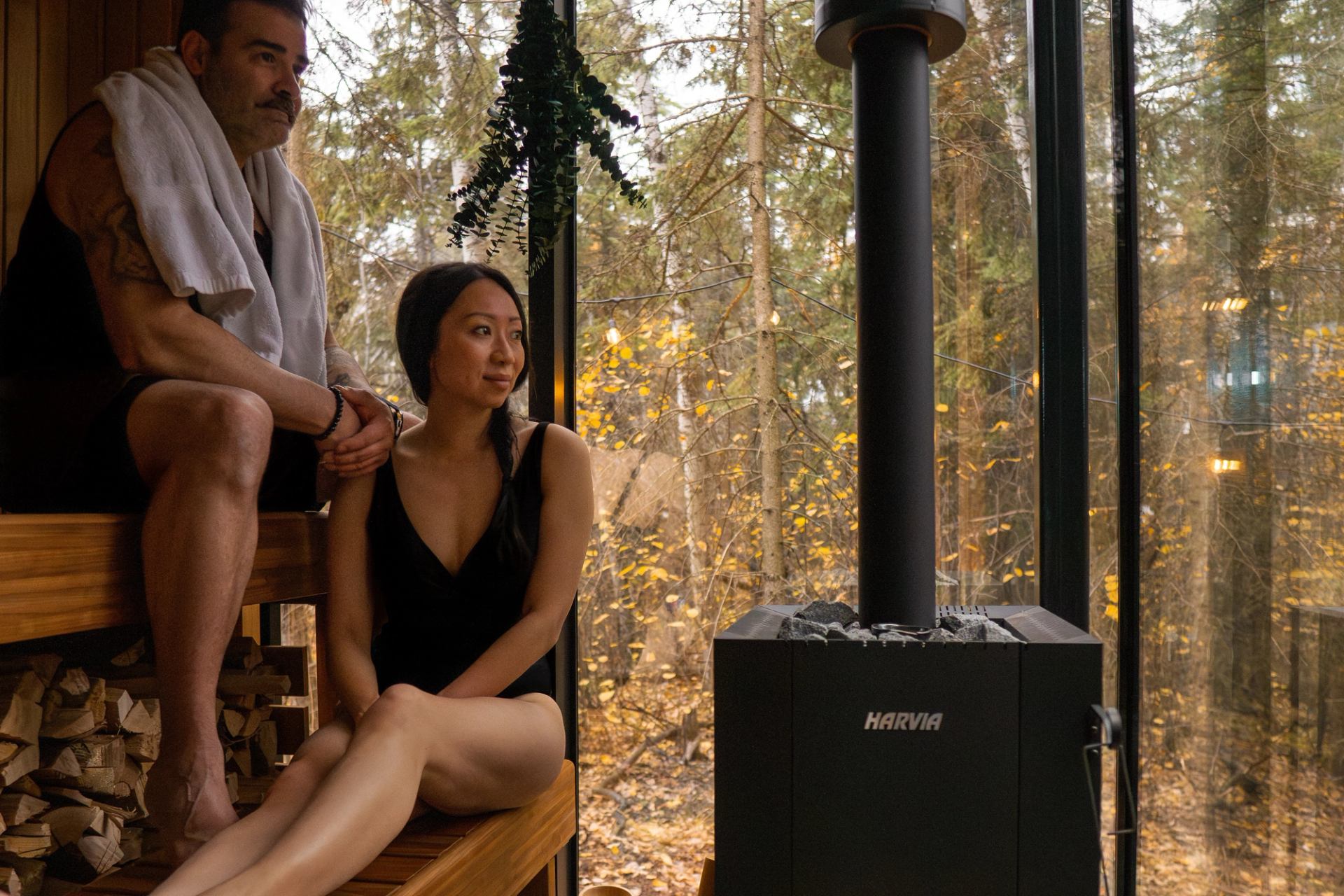 Two people lounge on the benches inside the mirrored sauna at Elk Island Retreat, with a glass wall revealing a view of the surrounding forest.