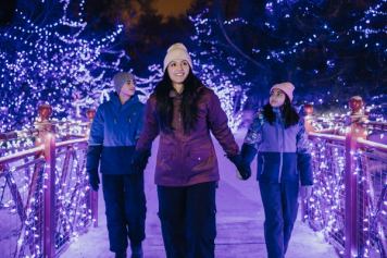 Three people walk on a snowy path lit by purple and white holiday lights at Calgary ZOOLIGHTS. The center person wears a maroon jacket and white beanie, holding hands with two others in blue jackets.