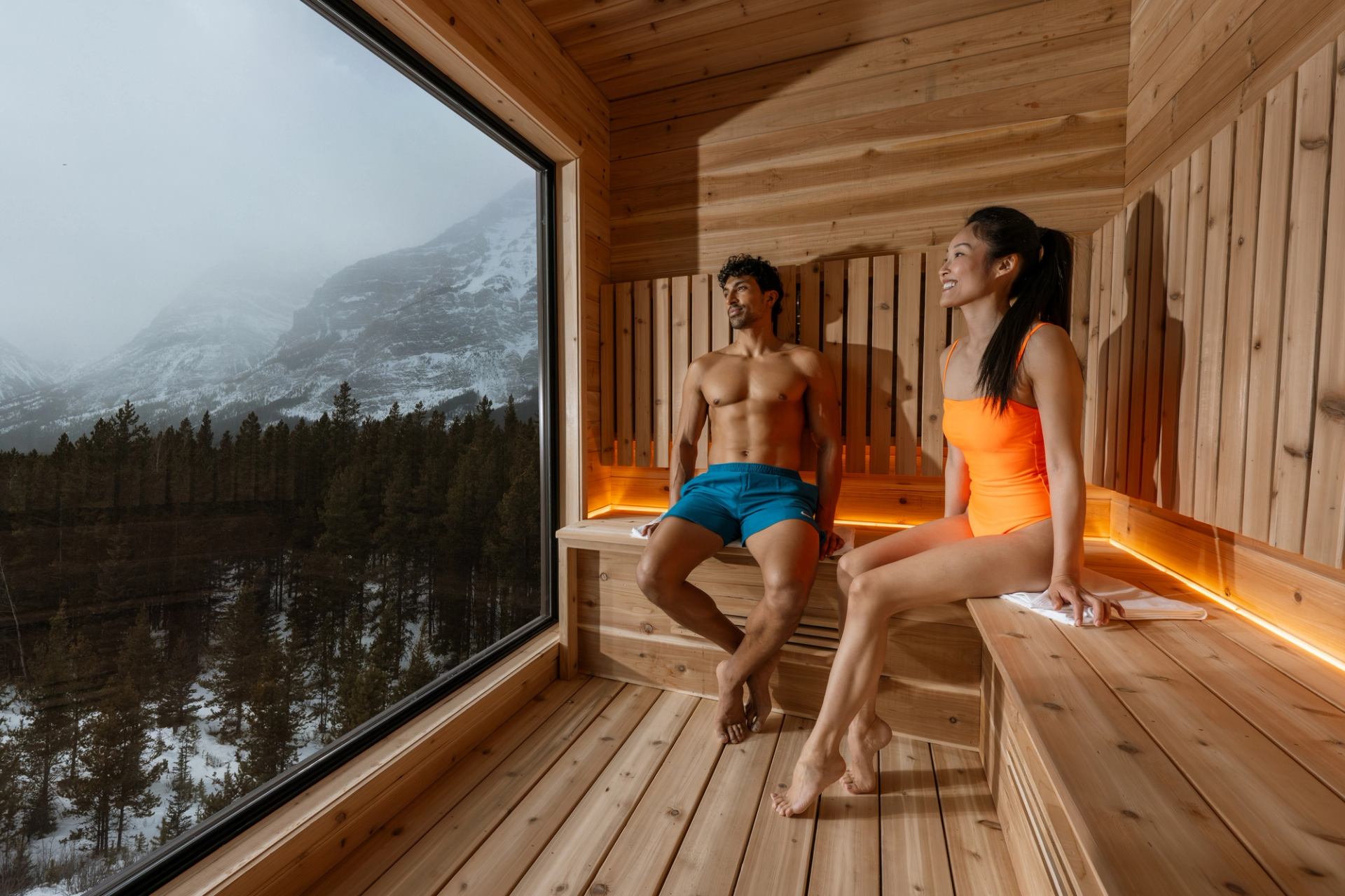 Two people relax in a sauna with views to the Rocky Mountains out a floor-to-ceiling window.