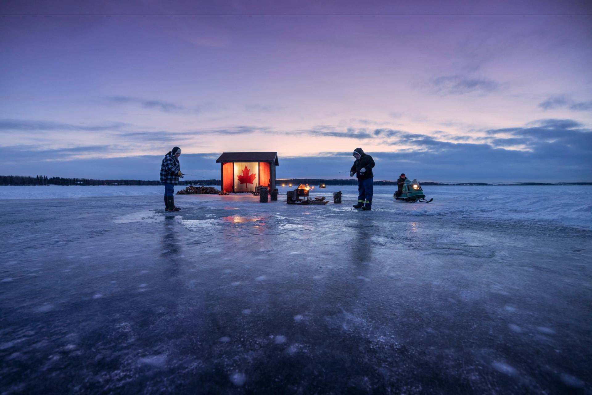 Two people ice fishing at sunset with an ice hut nearby.